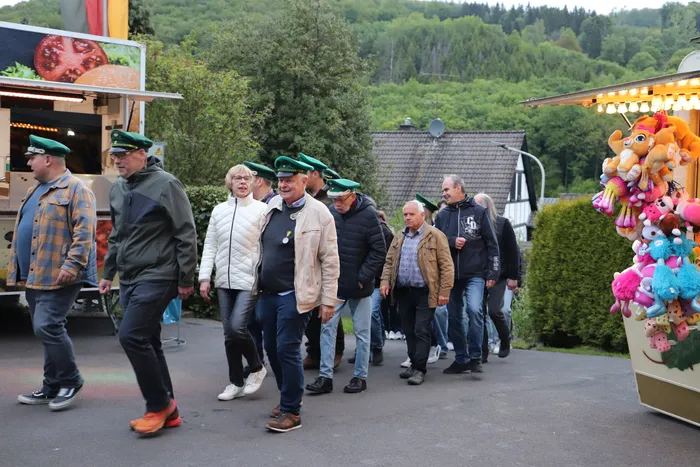 Eine Gruppe von Männern in Alltagskleidung und einigen mit grünen Schützenhüten geht auf einer asphaltierten Fläche an einem Imbisswagen und einem Spielstand vorbei.