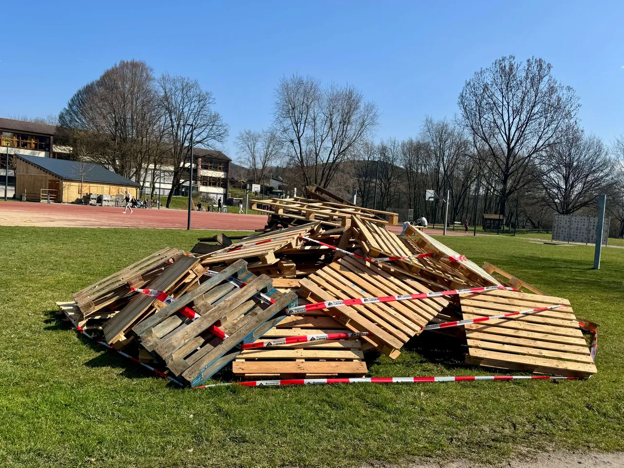 Ein großer Haufen Holzpaletten auf einer Wiese, umgeben von rotem Absperrband, im Hintergrund ein Sportplatz und Bäume.