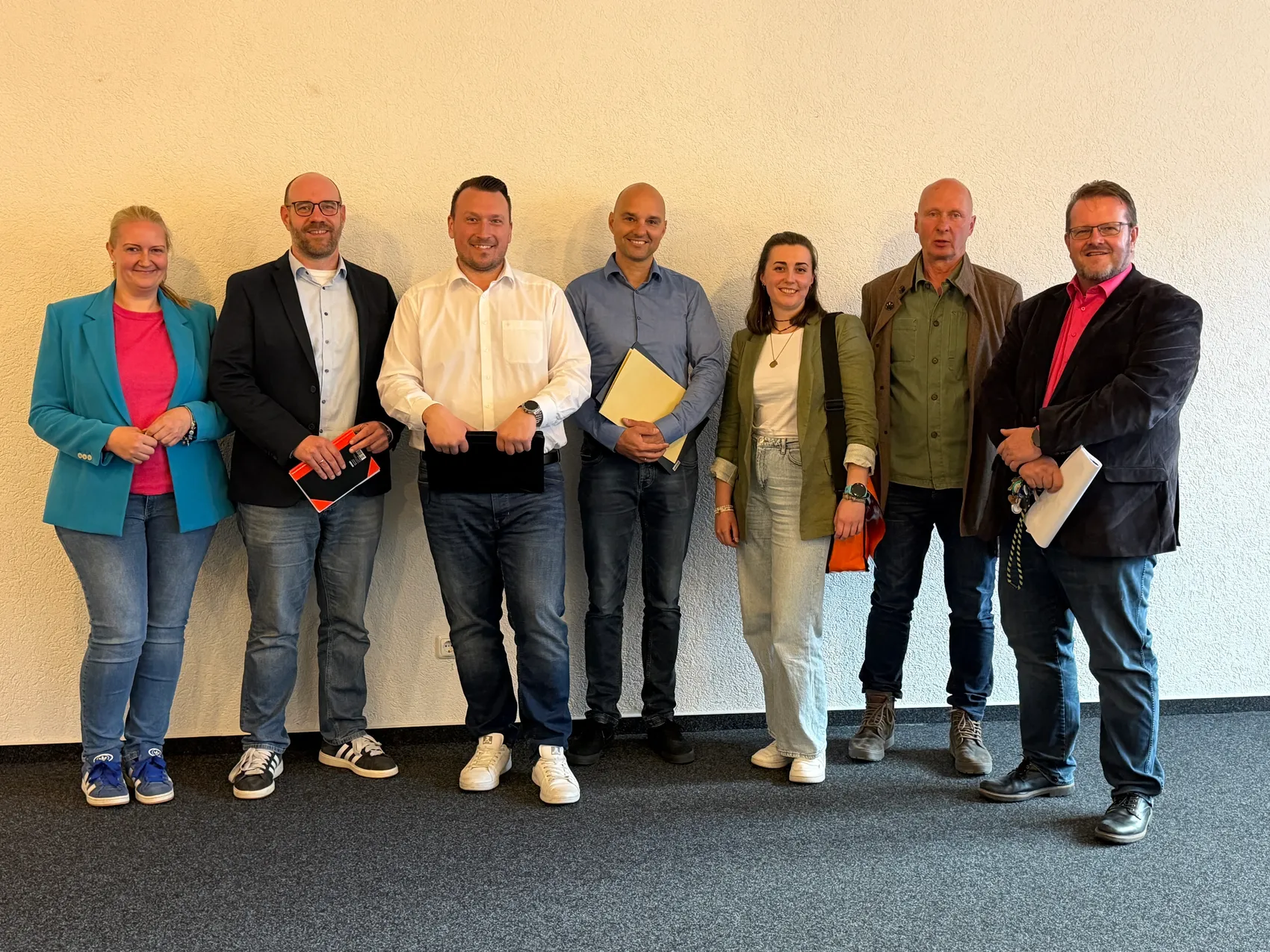 Sieben Personen stehen nebeneinander in einem hellen Raum. Sie halten teilweise Ordner oder Unterlagen in den Händen.