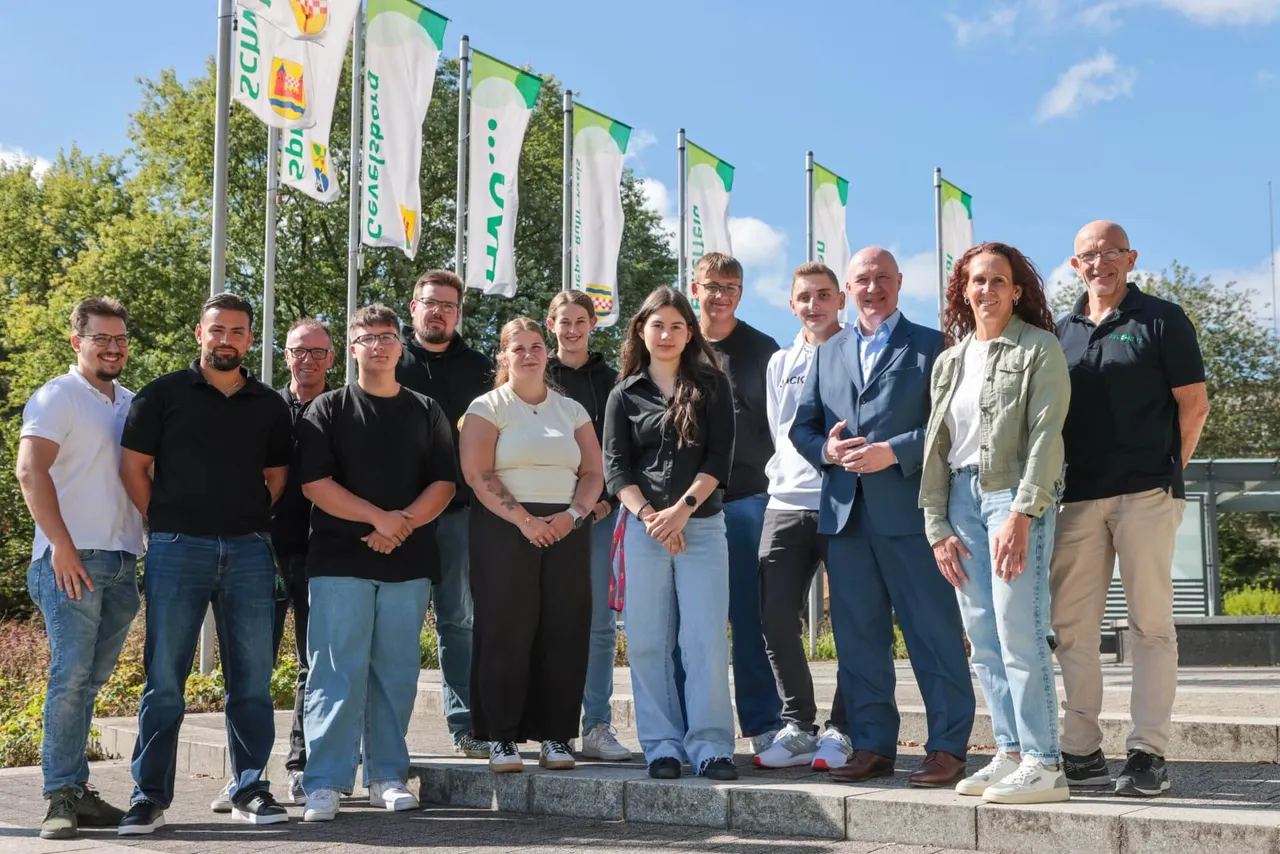 Eine Gruppe von AVU-Auszubildenden, Ausbildern und Führungskräften posiert für ein Gruppenfoto vor mehreren Fahnen im Freien.