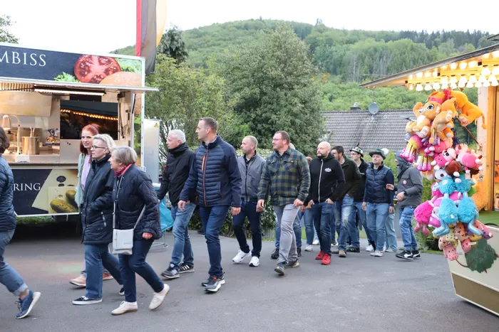 Menschen gehen an einem Imbissstand und einem Spielstand mit Stofftieren vorbei, im Hintergrund ein bewaldeter Hügel.