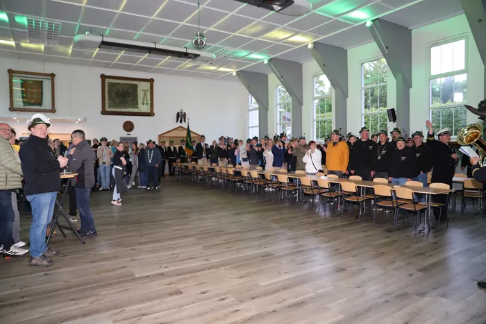 Große Menschengruppe in einem hellen Saal, Kimberly Walter mit Schützenhut und Orden im Mittelpunkt, rechts eine Person mit einer Posaune.