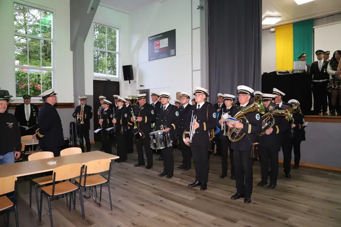 Blaskapelle in Uniformen spielt in einem hellen Raum, während im Hintergrund eine Frau auf einer kleinen Bühne steht.