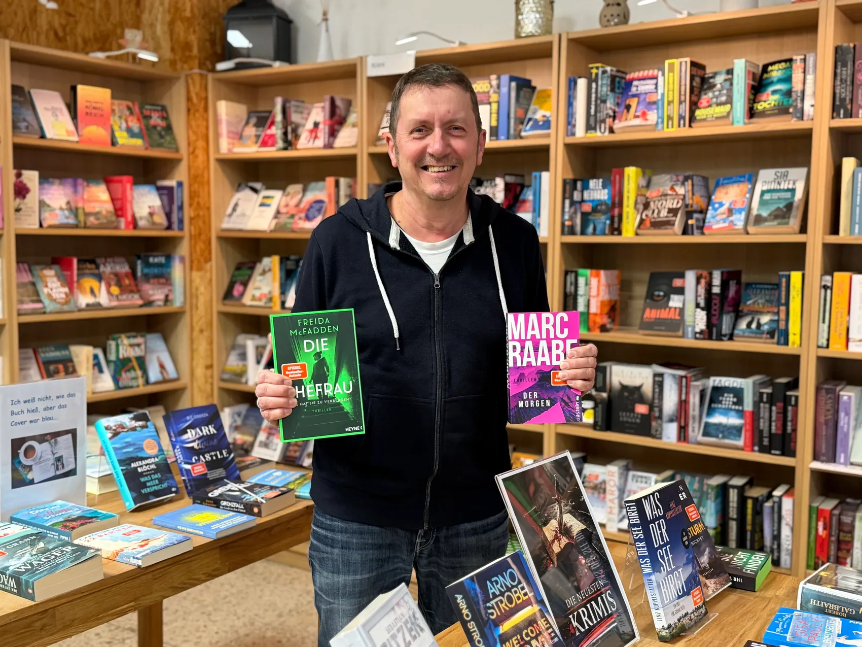 Martin Osthoff steht lächelnd in seinem Buchladen am Rathausplatz und hält ein Buch von Freida McFadden hoch.
