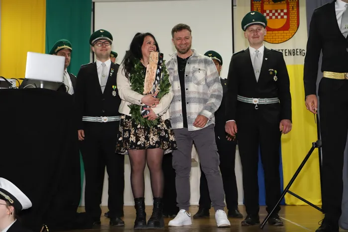 Kimberly Walter, die neue Schützenkönigin, hält einen Blumenstrauß und steht mit dem scheidenden Schützenkönig und weiteren Mitgliedern der Schützenbruderschaft auf einer Bühne. Im Hintergrund ist das Wappen von Landemert zu sehen.