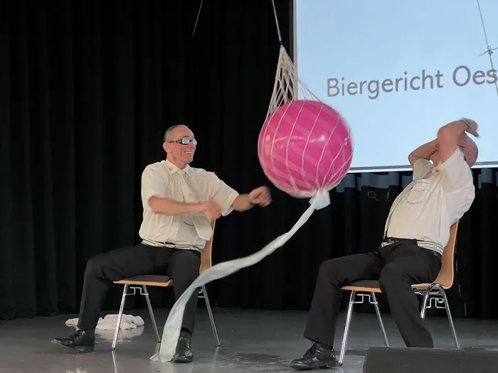 Zwei Männer sitzen auf Stühlen und halten an langen Bändern einen großen, pinken Ball hoch. Im Hintergrund ist eine Projektion mit dem Text „Biergericht Oest“ zu sehen.