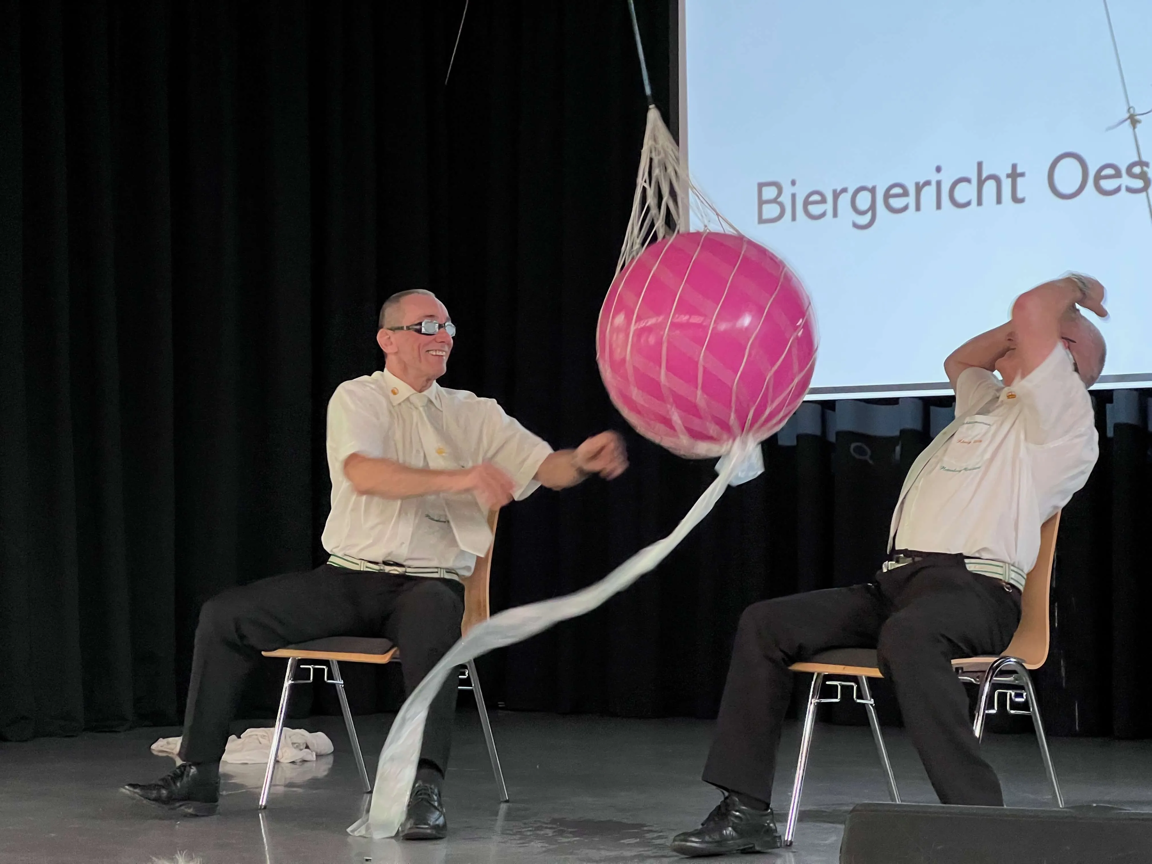 Zwei Männer sitzen auf Stühlen und halten an langen Bändern einen großen, pinken Ball hoch. Im Hintergrund ist eine Projektion mit dem Text „Biergericht Oest“ zu sehen.