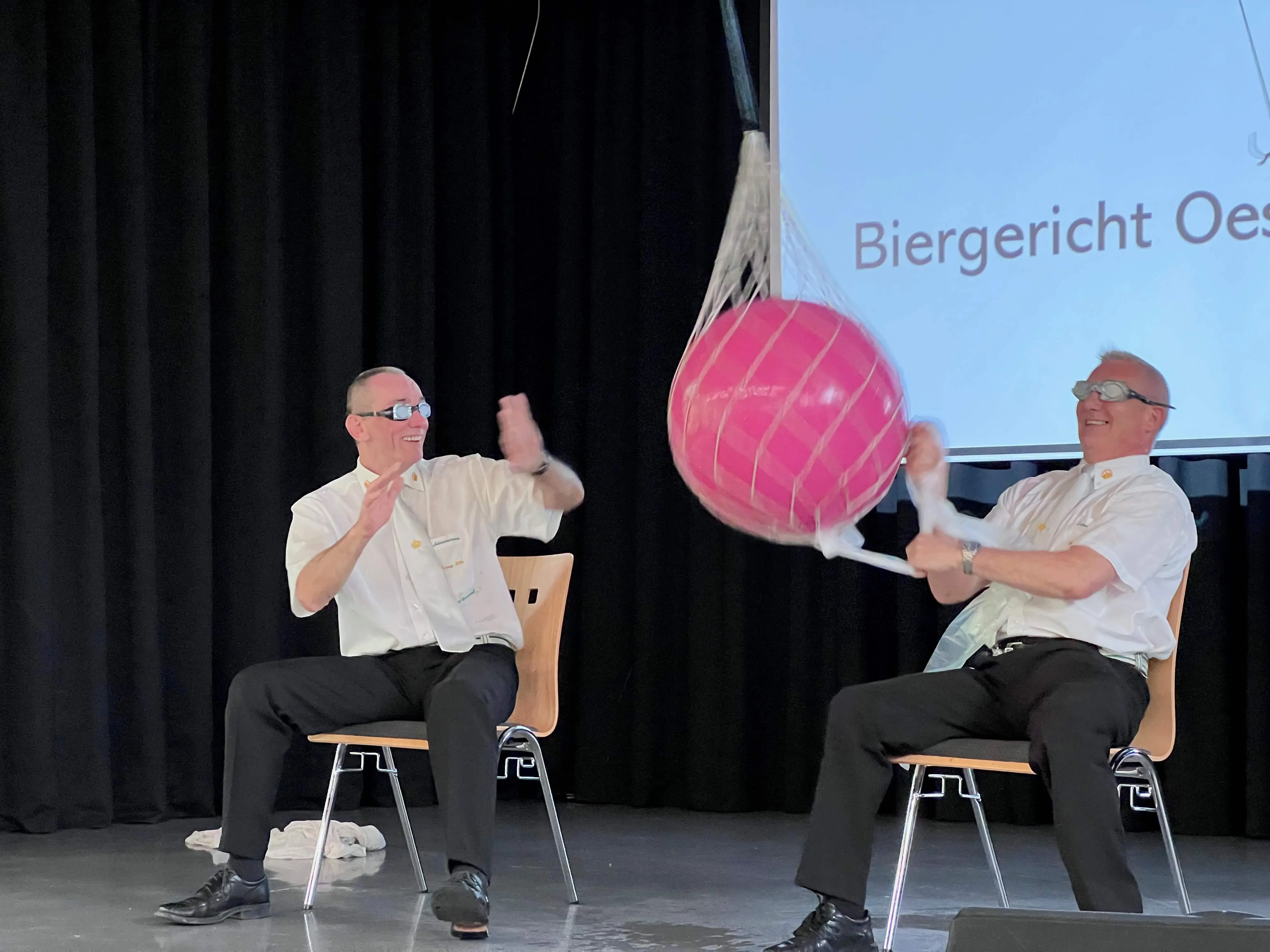 Zwei Männer sitzen auf Stühlen auf einer Bühne und werfen sich einen großen, pinkfarbenen Ball zu. Im Hintergrund eine Projektionsfläche mit dem Text „Biergericht Oestertal“.