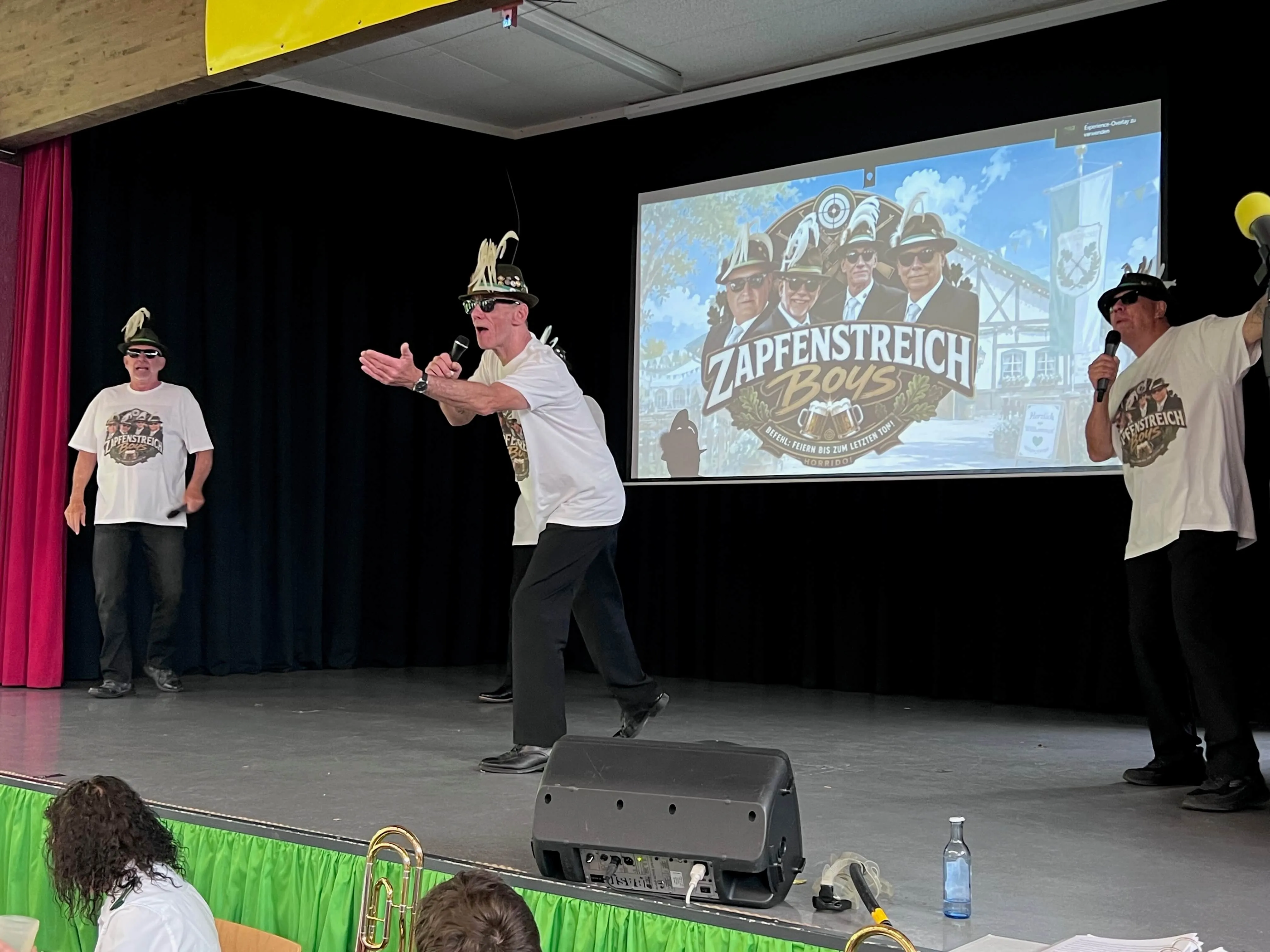 Drei Männer in traditioneller Tracht stehen auf einer Bühne vor einem großen Bildschirm mit der Aufschrift „Zapfenstreich Boys“. Der Mann in der Mitte gestikuliert lebhaft, während die anderen beiden ihn beobachten.