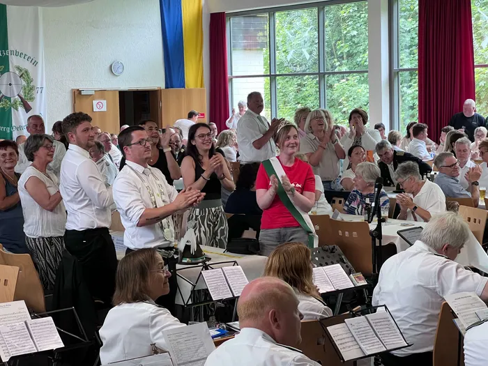 Applaus im Raum, in dem ein Blasorchester spielt. Im Vordergrund eine Frau mit Blumenkranz und rotem Oberteil, umgeben von applaudierenden Menschen unterschiedlichen Alters.