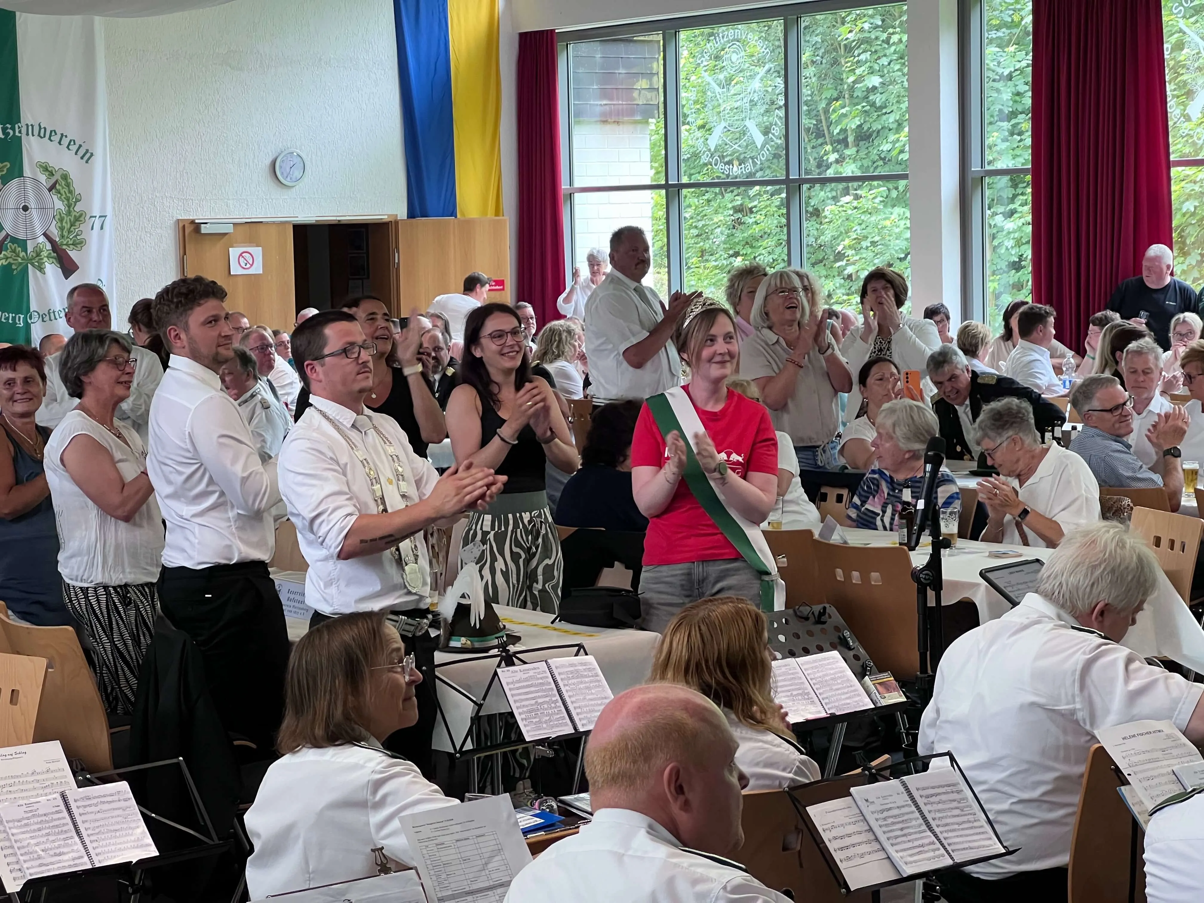 Applaus im Raum, in dem ein Blasorchester spielt. Im Vordergrund eine Frau mit Blumenkranz und rotem Oberteil, umgeben von applaudierenden Menschen unterschiedlichen Alters.