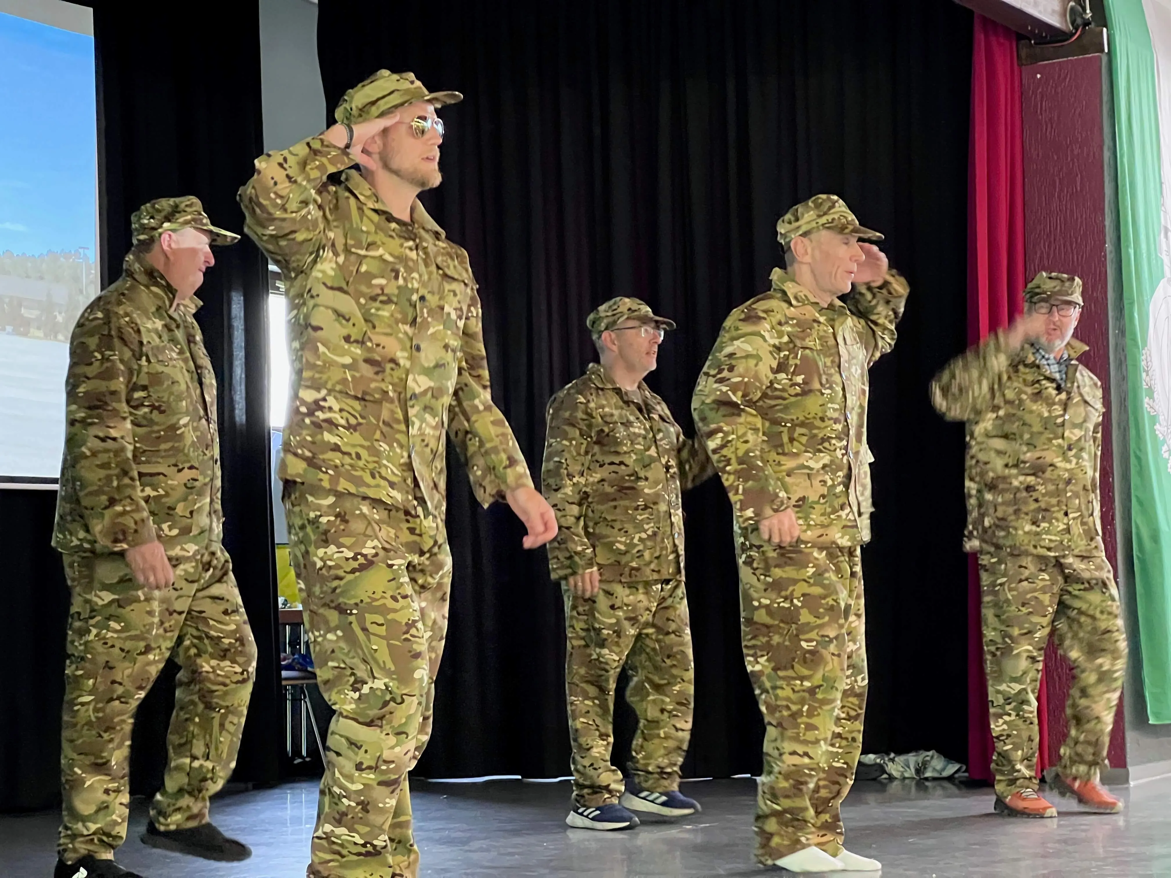Fünf Personen in Tarnuniformen stehen auf einer Bühne und salutieren mit der rechten Hand. Im Hintergrund ist ein roter Vorhang und eine Projektionsfläche zu sehen.