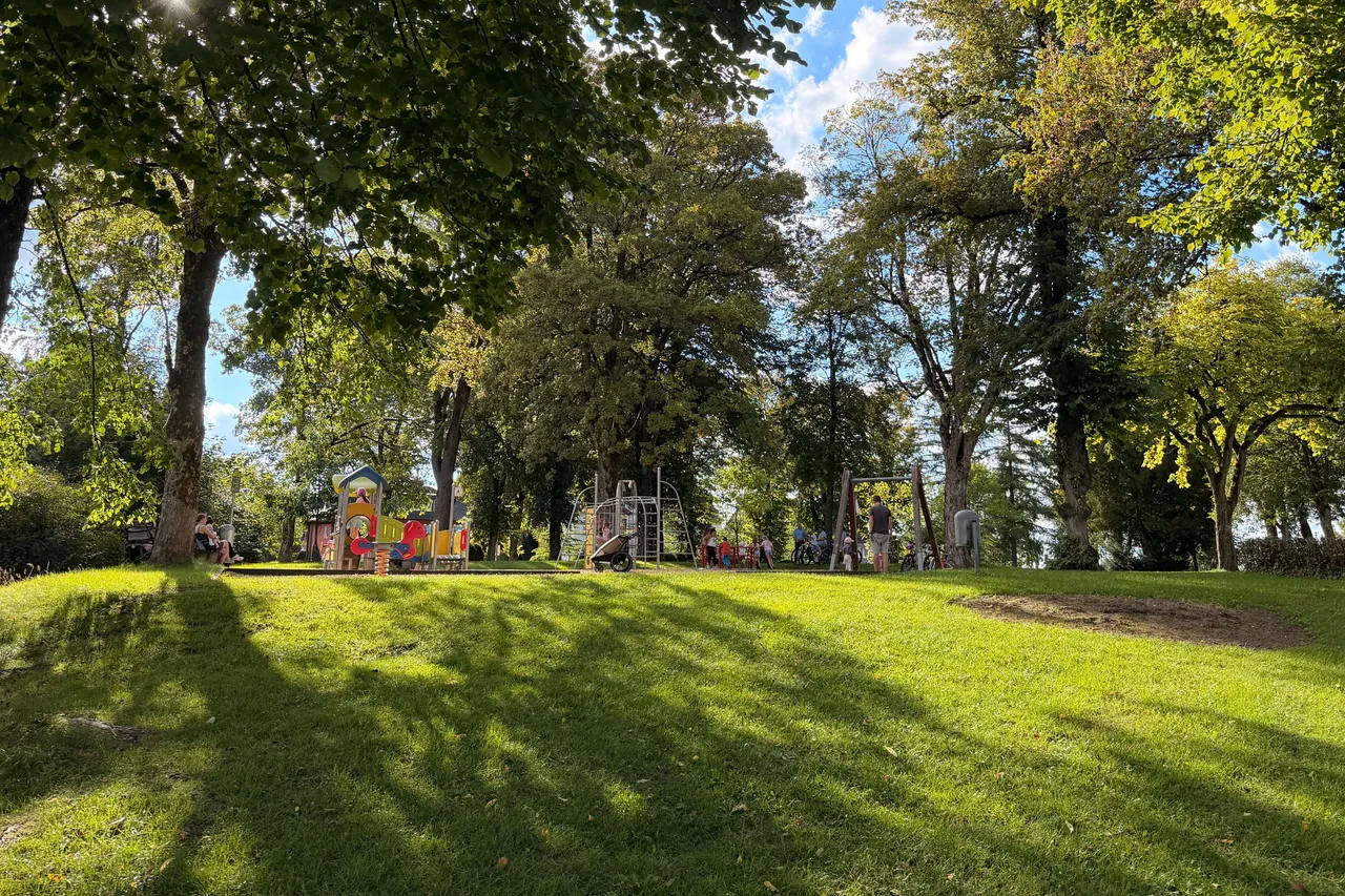 Sonniger Spielplatz im Park mit Rasenflächen, großen Bäumen und Menschen an verschiedenen Spielgeräten.