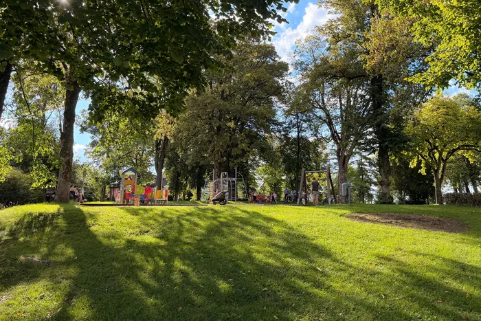 Sonniger Spielplatz im Park mit Rasenflächen, großen Bäumen und Menschen an verschiedenen Spielgeräten.
