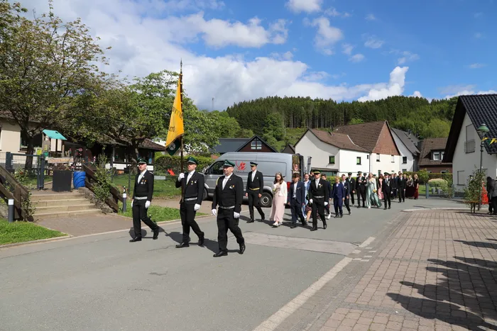 Eine Gruppe uniformierter Männer und Frauen marschiert auf einer Straße, vornewein Fahrenträger. Im Hintergrund sind Häuser und grüne Hügel zu sehen.