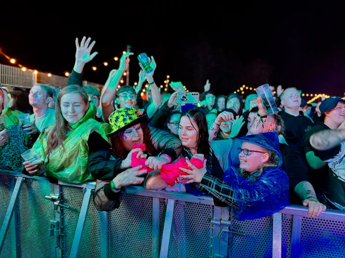 Eine jubelnde Menschenmenge hinter einer Metallabsperrung bei einem Musikfestival bei Nacht, teilweise in bunten Regenponchos.