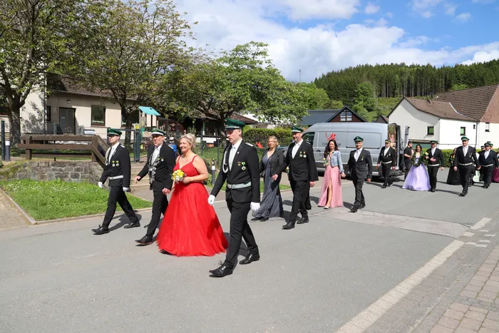 Eine Gruppe von Menschen in festlicher Tracht schreitet auf einer Straße. Eine Frau in einem roten Ballkleid trägt einen Blumenstrauß.