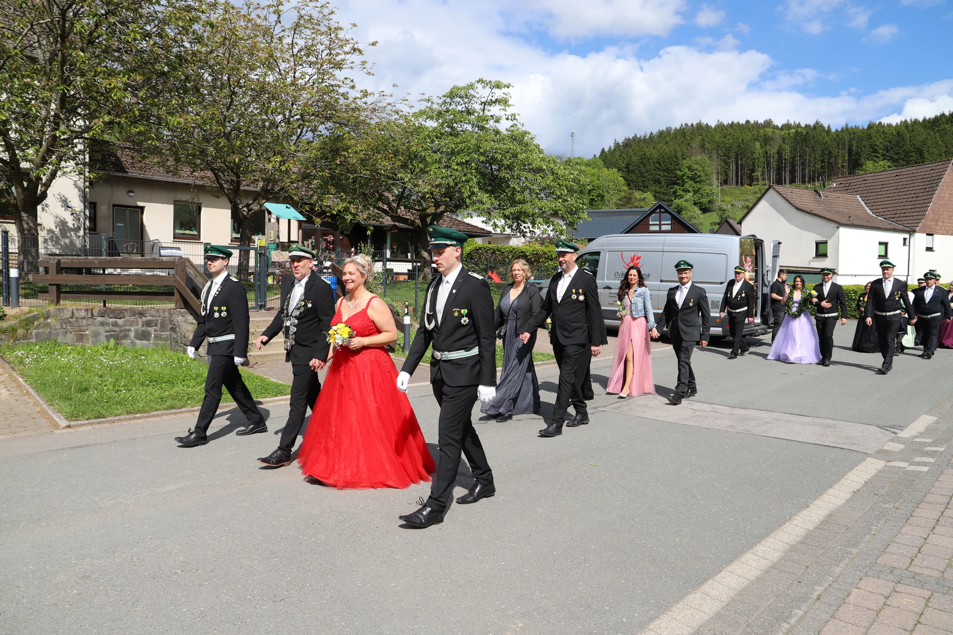 Eine Gruppe von Menschen in festlicher Tracht schreitet auf einer Straße. Eine Frau in einem roten Ballkleid trägt einen Blumenstrauß.