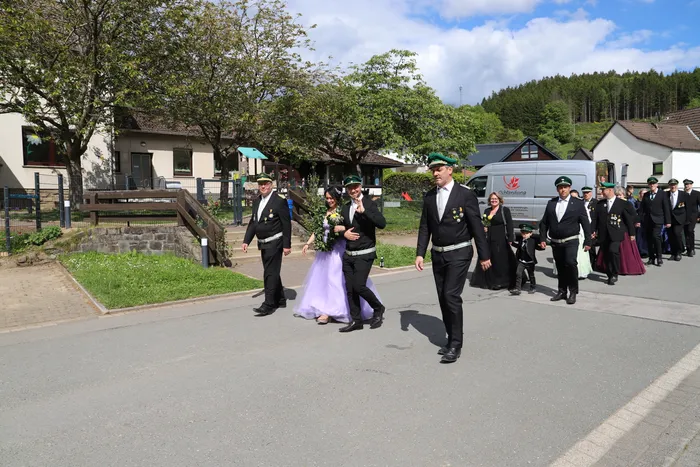 Eine Gruppe von Menschen in traditionellen Uniformen schreitet auf einem Weg zu, eine Frau trägt ein lilafarbenes Kleid und einen Blumenkranz.