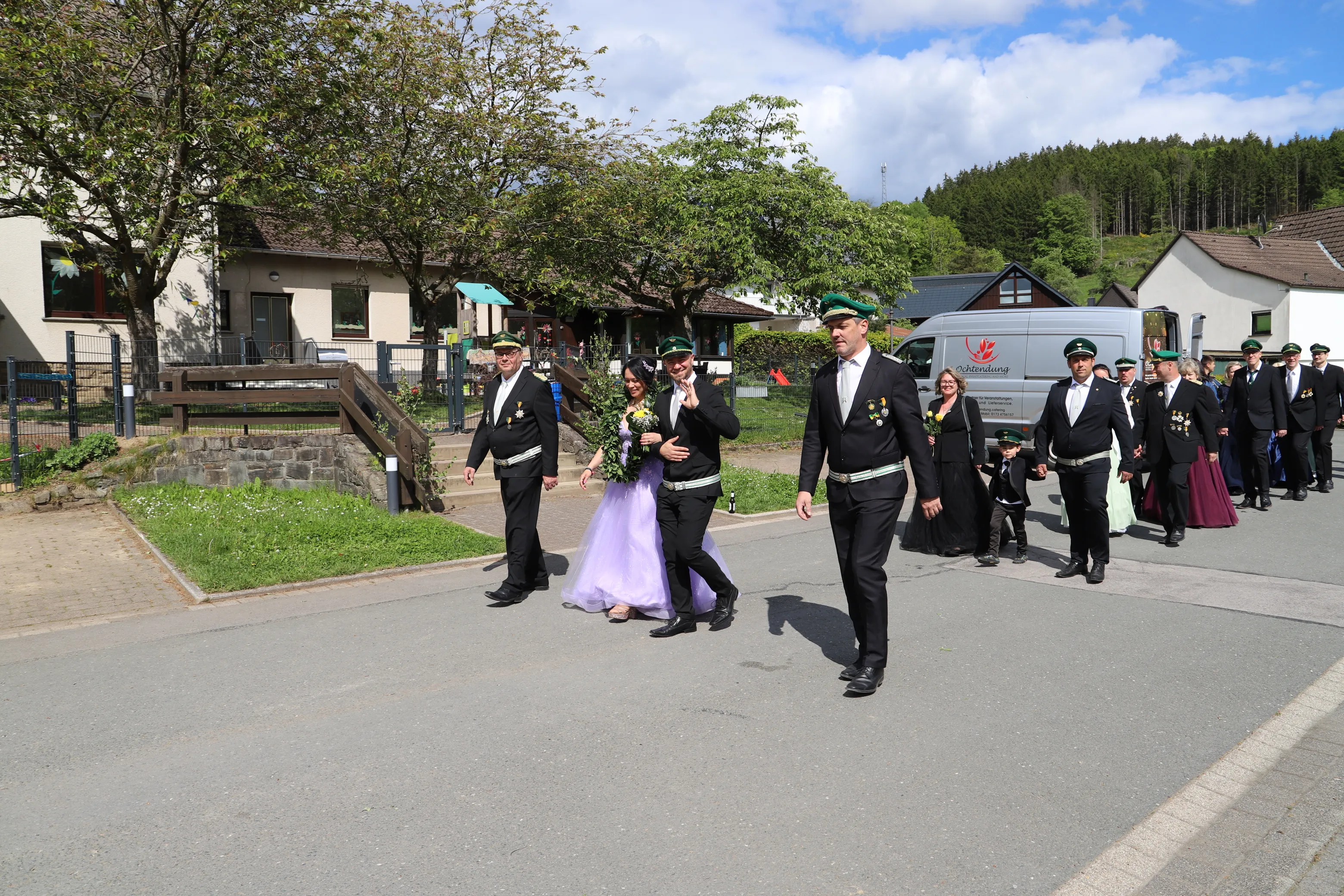 Eine Gruppe von Menschen in traditionellen Uniformen schreitet auf einem Weg zu, eine Frau trägt ein lilafarbenes Kleid und einen Blumenkranz.