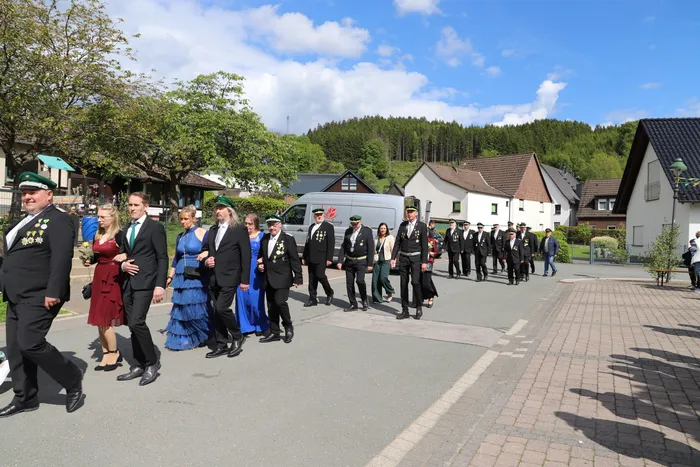 Eine Gruppe von Menschen in festlicher Tracht schreitet auf einem gepflasterten Platz vor einer ländlichen Kulisse. Viele tragen grüne oder schwarze Anzüge mit Abzeichen und Hüten.