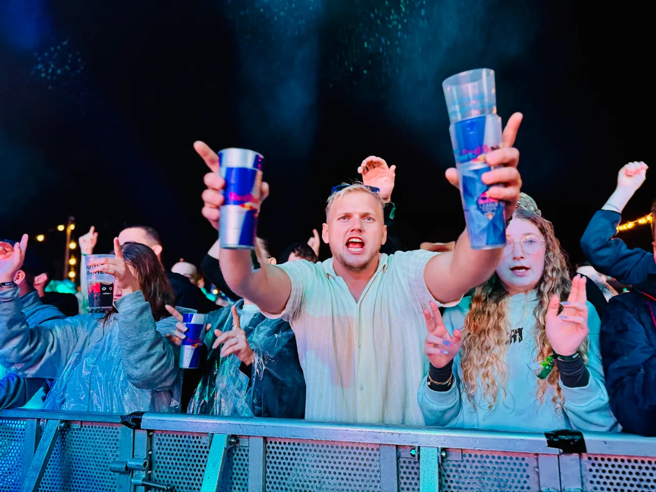 Ein jubelnder Mann hält zwei blaue Becher hoch in einer Menschenmenge bei einem Festival.