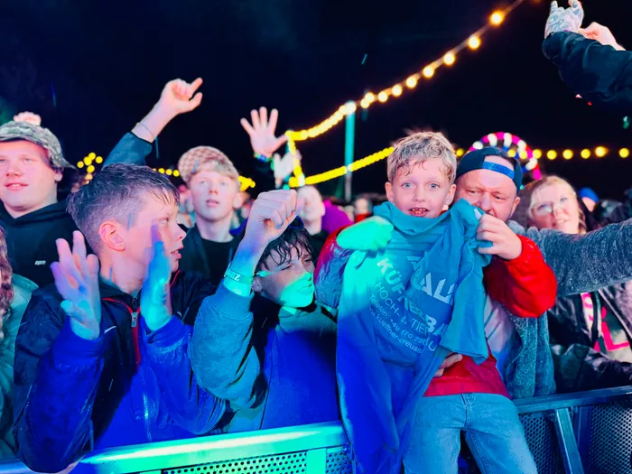 Eine jubelnde Menschenmenge bei einem nächtlichen Festival, beleuchtet von bunten Lichtern und Lichterketten.