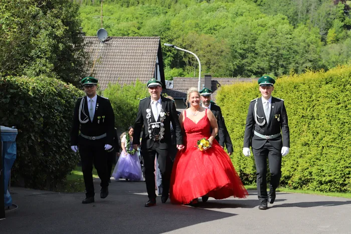 Eine Frau in rotem Ballkleid schreitet lächelnd zwischen vier Männern in Uniformen und einer jungen Blumenjungfrau auf einem Weg entlang.