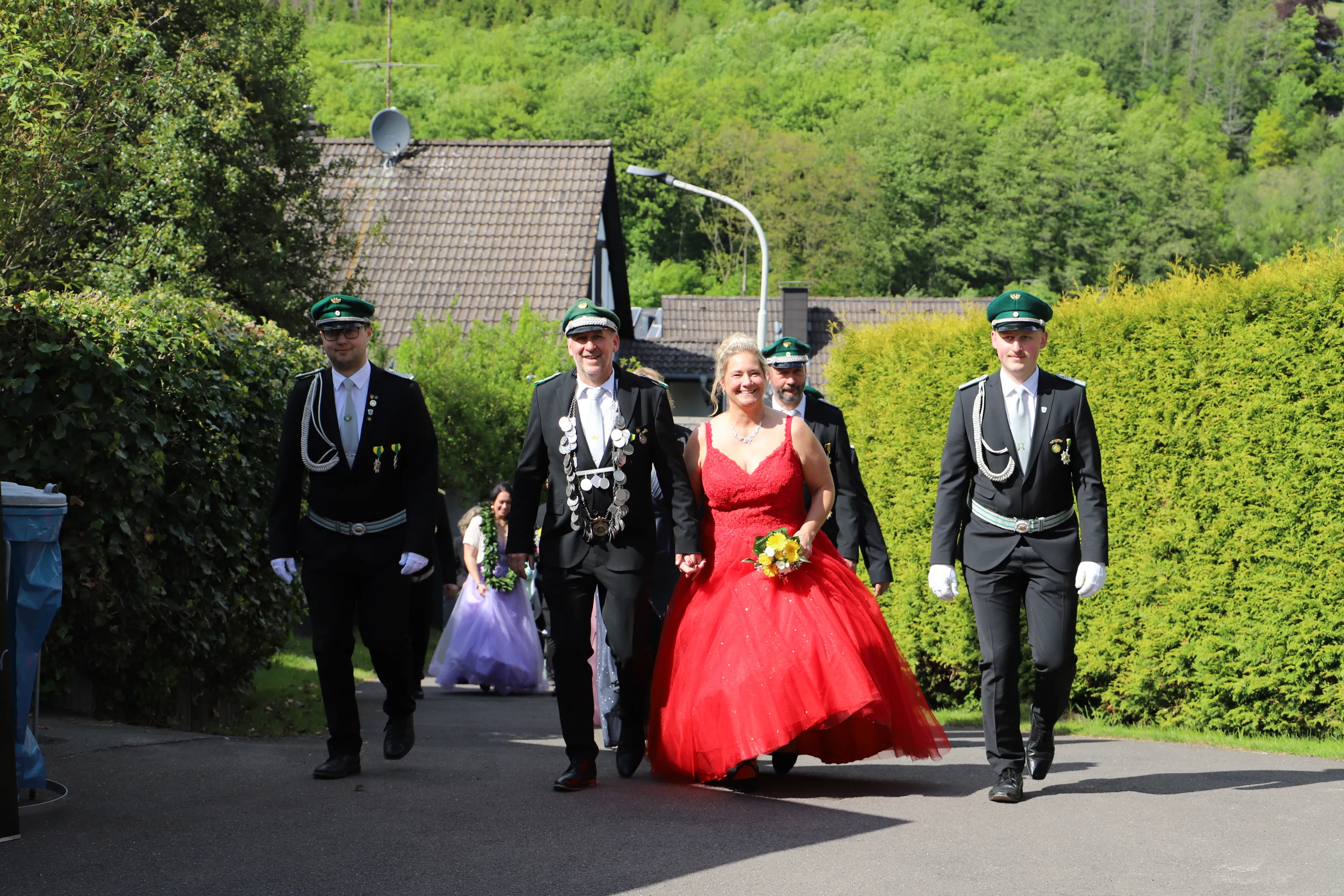 Eine Frau in rotem Ballkleid schreitet lächelnd zwischen vier Männern in Uniformen und einer jungen Blumenjungfrau auf einem Weg entlang.