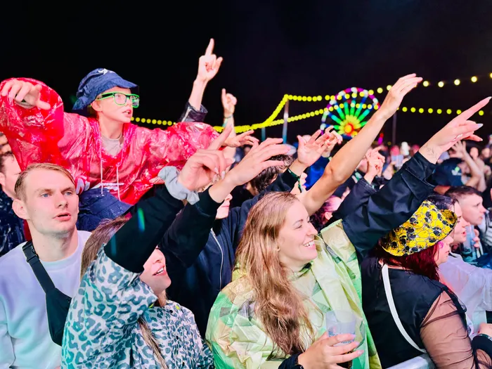 Eine jubelnde Menschenmenge mit erhobenen Händen bei einem nächtlichen Open-Air-Event mit bunten Lichtern im Hintergrund.