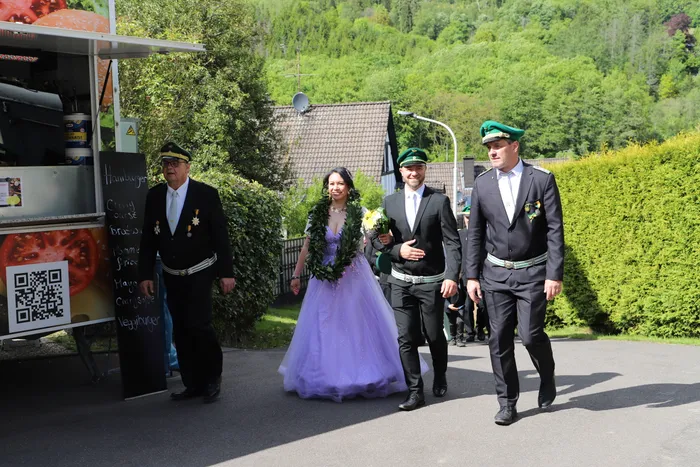 Vier Personen in Festkleidung gehen an einem Imbisswagen vorbei. Die Frau trägt ein lilafarbenes Kleid und eine Blütengirlande, die Männer Uniformen mit Zylinder und Abzeichen.