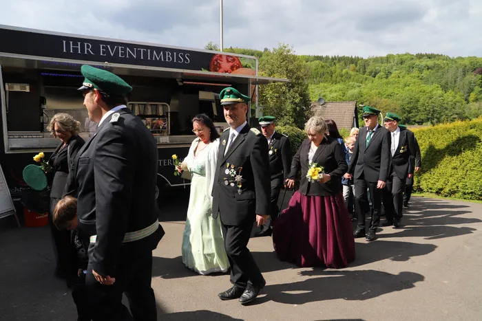 Eine Gruppe von Menschen in festlicher Tracht geht an einem Imbisswagen vorbei. Einige tragen grüne Hüte und Uniformen, andere sind in Abendkleidern gekleidet.