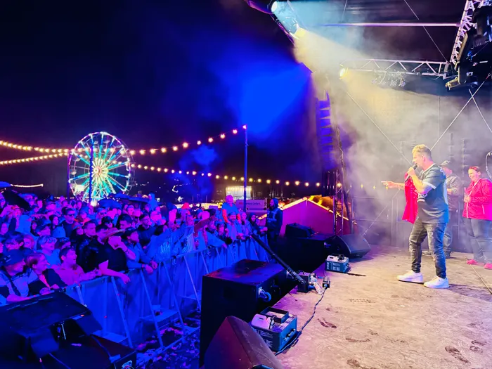 Almklausi performt auf einer Bühne vor einer großen Menschenmenge. Im Hintergrund sind ein Riesenrad und Lichterketten zu sehen.