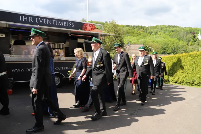 Eine Gruppe von Menschen in dunklen Anzügen und einigen mit grünen Schützenhüten geht an einem Foodtruck vorbei, der mit „Ihr Eventimbiss“ beworben wird. Im Hintergrund sind Bäume und Häuser zu sehen.