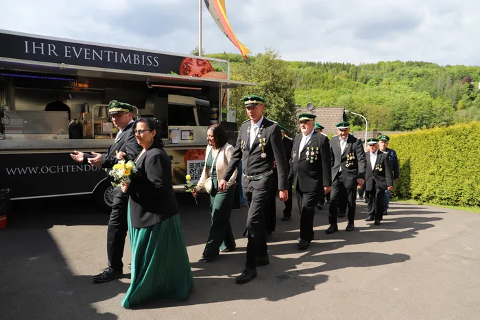 Eine Gruppe von Menschen in dunklen Uniformen und traditioneller Kleidung geht an einem Foodtruck vorbei, im Hintergrund ein bewaldeter Hang und die deutsche Flagge.