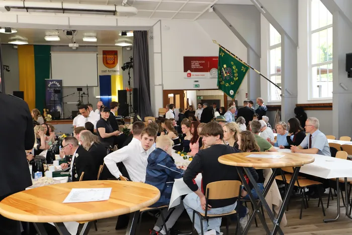 Menschen sitzen an Tischen in einem hellen Raum und unterhalten sich. Im Hintergrund sind Wappen und eine grüne Flagge zu sehen.