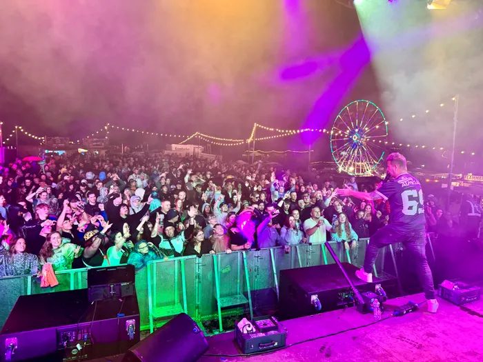 Almklausi performt auf einer Bühne vor einer großen Menschenmenge bei einem nächtlichen Festival mit bunten Lichtern und einem Riesenrad im Hintergrund.