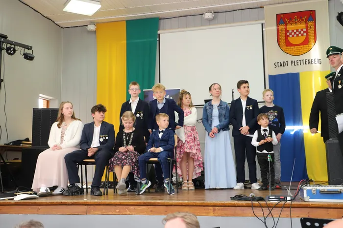 Eine Gruppe von Kindern und Jugendlichen in festlicher Kleidung sitzt auf Stühlen auf einer Bühne. Im Hintergrund ist das Wappen der Stadt Plettenberg zu sehen, daneben steht ein Mann in Uniform.