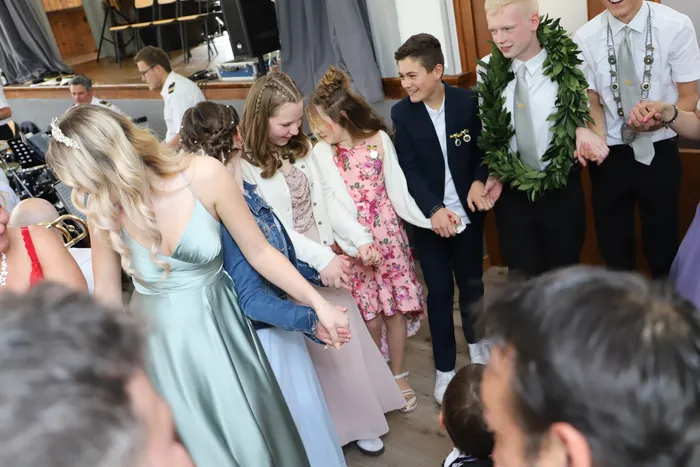 Eine Gruppe von Jugendlichen und jungen Erwachsenen hält sich an den Händen und tanzt im Kreis, einige tragen festliche Kleidung und Blumenschmuck.