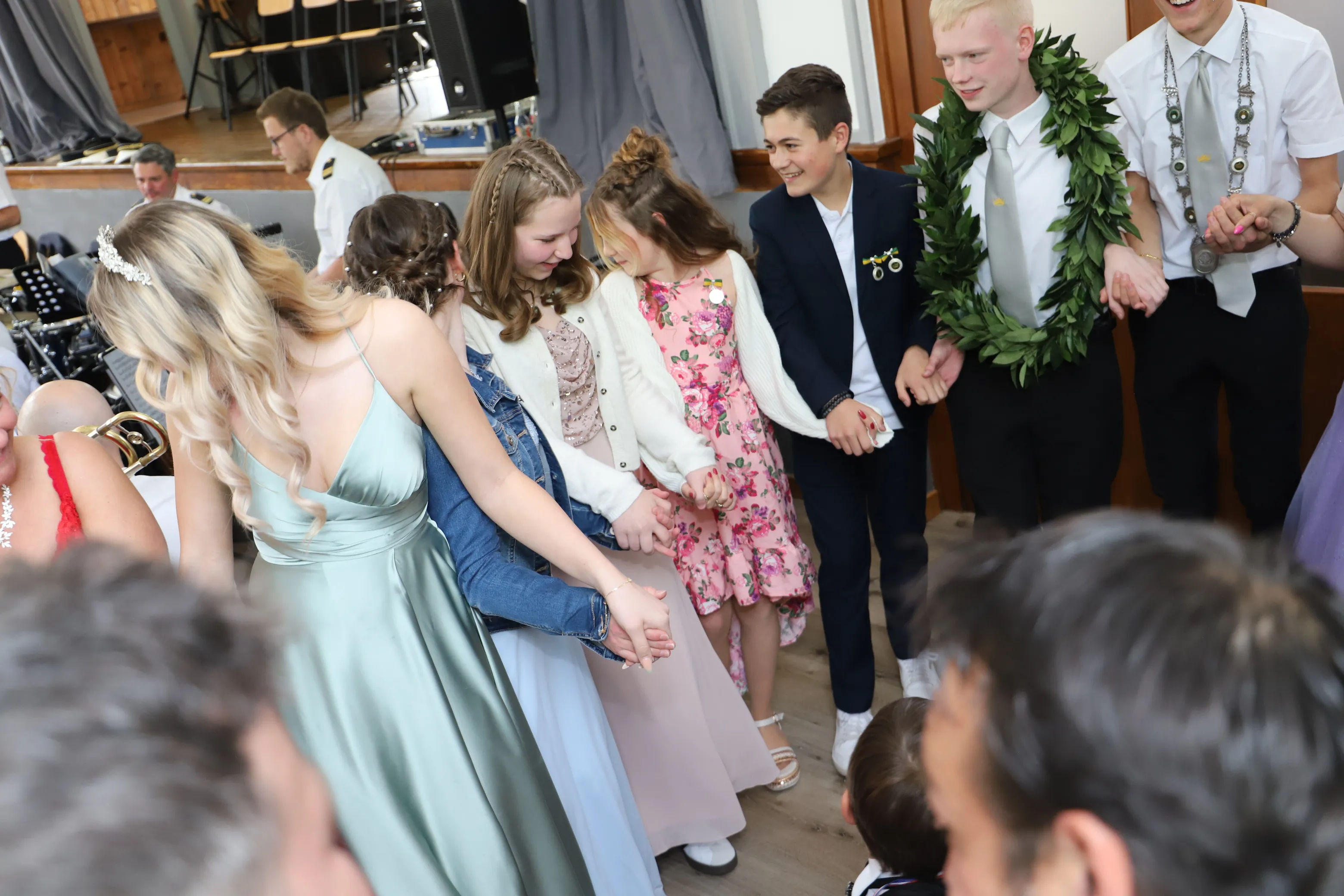 Eine Gruppe von Jugendlichen und jungen Erwachsenen hält sich an den Händen und tanzt im Kreis, einige tragen festliche Kleidung und Blumenschmuck.