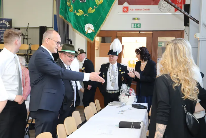 Zwei Männer im Anzug und ein Mann in Schützenuniform geben sich die Hände. Im Hintergrund stehen weitere Personen und ein Tisch mit Infomaterialien.
