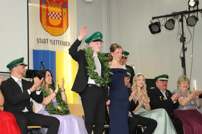 Ein Teenager mit Schützenhut und Lorbeerkranz steht auf einer Bühne neben einem Mädchen in Abendkleid, beide lächeln in die Kamera. Im Hintergrund applaudieren Menschen und ist das Stadtwappen von Plettenberg zu sehen.