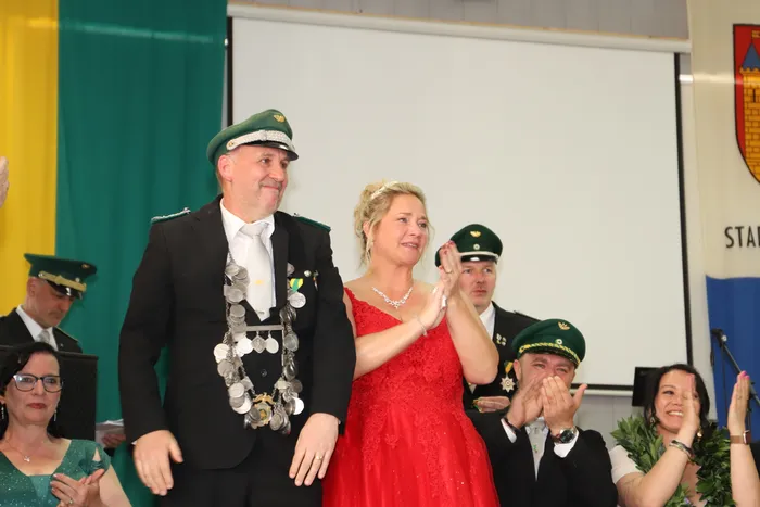 Ein Mann in Schützenuniform und eine Frau in rotem Kleid stehen auf einer Bühne und werden von anderen Personen bejubelt. Im Hintergrund ist das Wappen der Stadt zu sehen.
