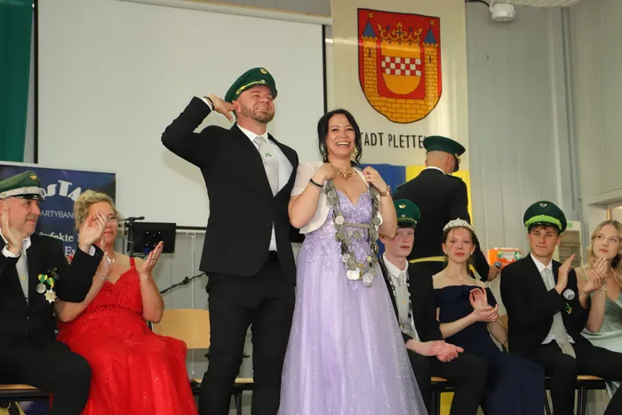 Ein Mann und eine Frau in traditioneller Schützenkleidung stehen auf einer Bühne und werden von anderen Mitgliedern des Schützenvereins Landemert gefeiert. Im Hintergrund ist das Wappen der Stadt Plettenberg zu sehen.