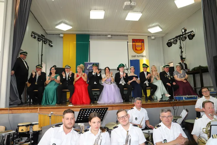 Festlich gekleidete Mitglieder des Schützenvereins Landemert sitzen auf einer Bühne, während eine Blaskapelle im Vordergrund spielt. Im Hintergrund ist das Stadtwappen von Plettenberg zu sehen.