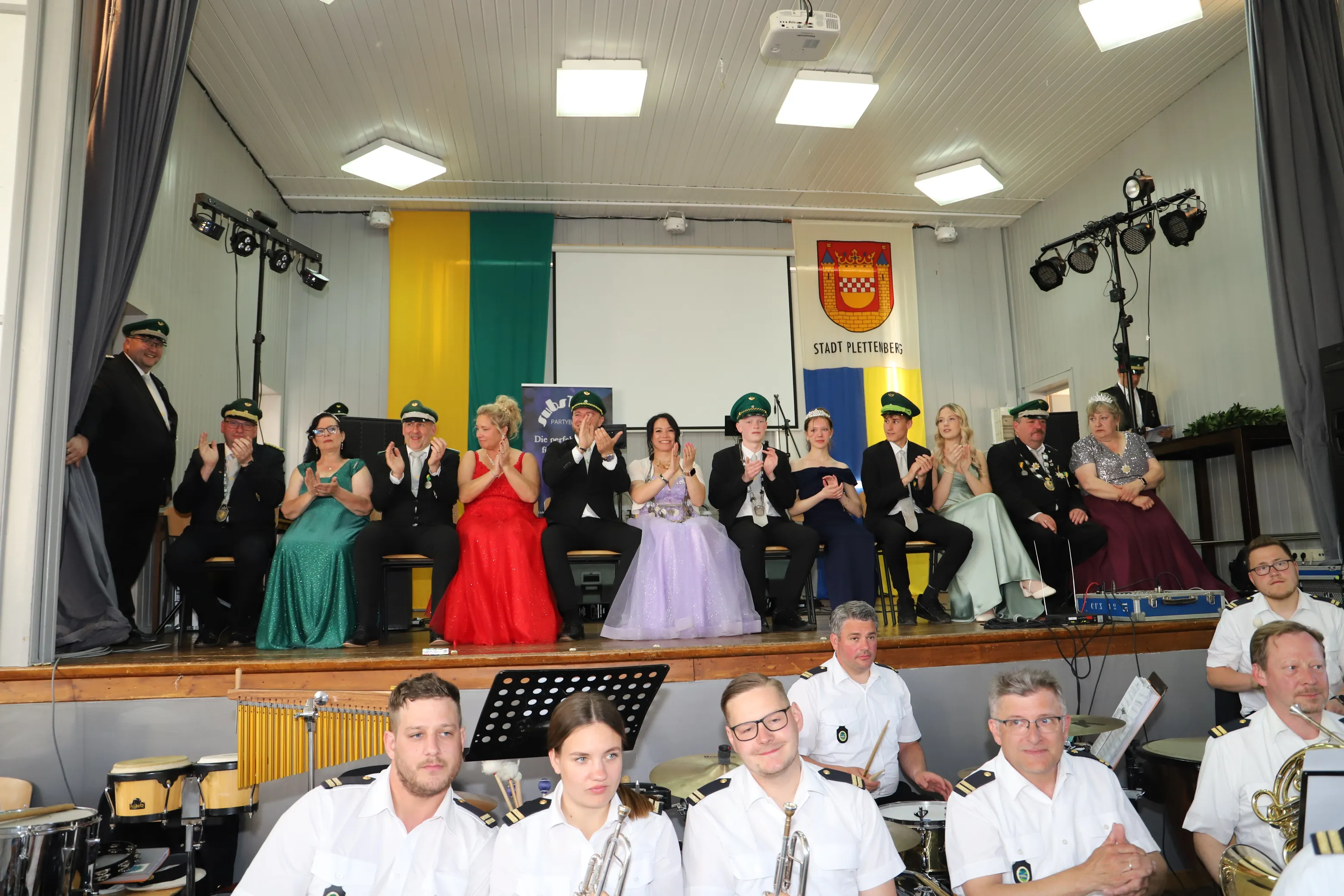 Festlich gekleidete Mitglieder des Schützenvereins Landemert sitzen auf einer Bühne, während eine Blaskapelle im Vordergrund spielt. Im Hintergrund ist das Stadtwappen von Plettenberg zu sehen.