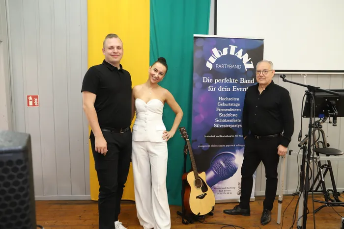 Drei Musiker, ein Mann und eine Frau im weißen Outfit, stehen vor einem Banner der Partyband „Substanz“ und einem Musikinstrument. Im Hintergrund ist Bühnentechnik zu sehen.