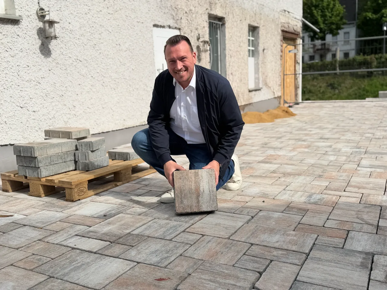 Bürgermeister Christian Breddermann kauert auf dem gepflasterten Hof des Hotels zur Post und hält einen Pflasterstein in der Hand. Im Hintergrund sind weitere Pflastersteine auf einer Palette und das Gebäude zu sehen.