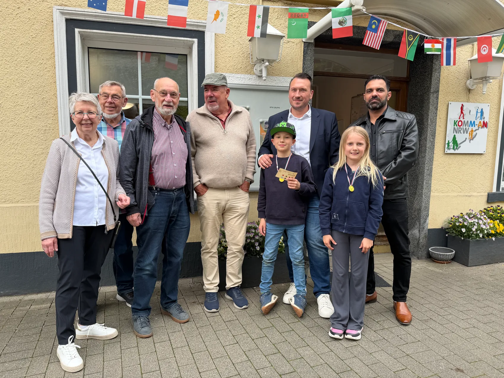 Gruppe von sieben Personen vor dem Westfälischen Hof in Schalksmühle, darunter ein Kind mit einer Medaille. Im Hintergrund sind Flaggen zu sehen.