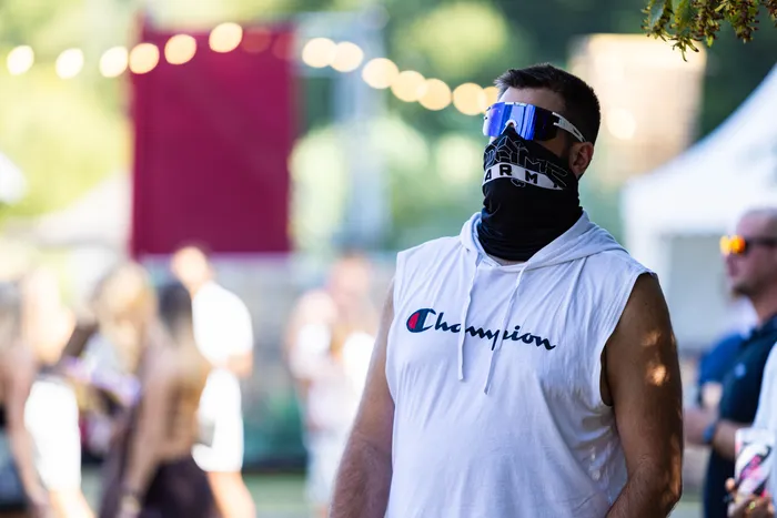 Ein Mann mit blauer Sonnenbrille, schwarzer Maske und weißem, ärmellosem Champion-Hoodie vor einem unscharfen Festivalgelände.